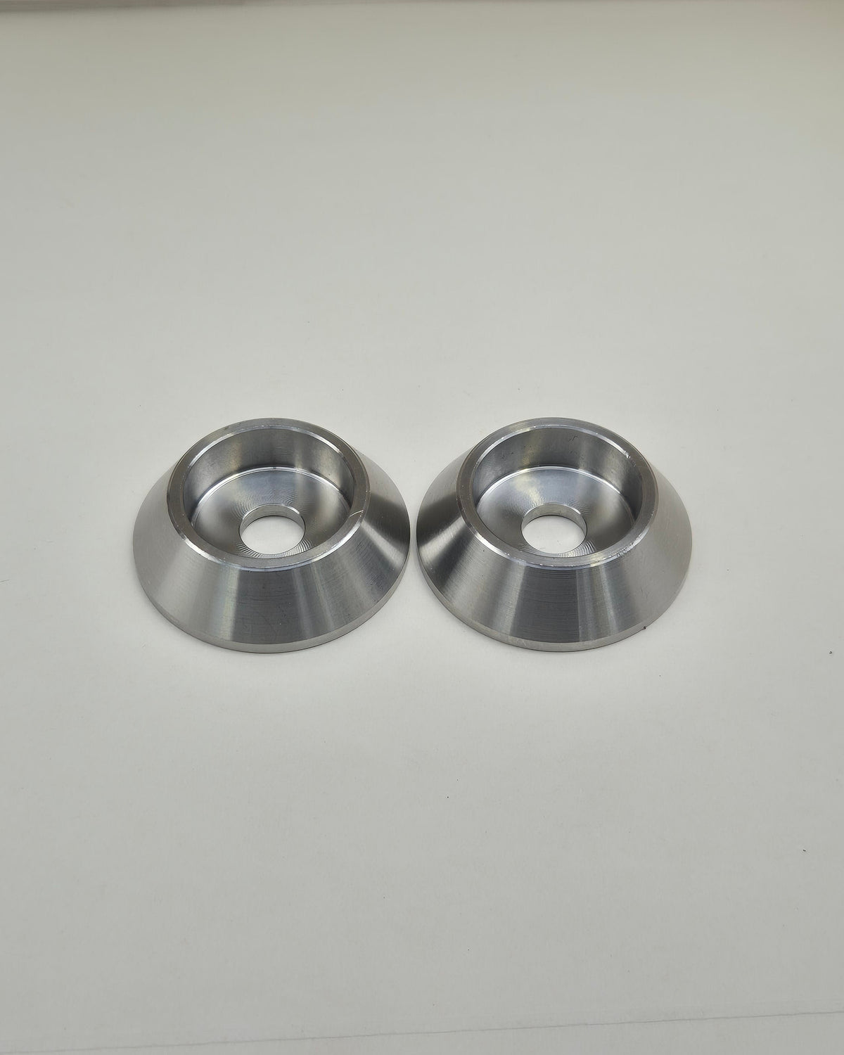 Two machined bolt head protectors with tapered outer walls and recessed centres on a light background